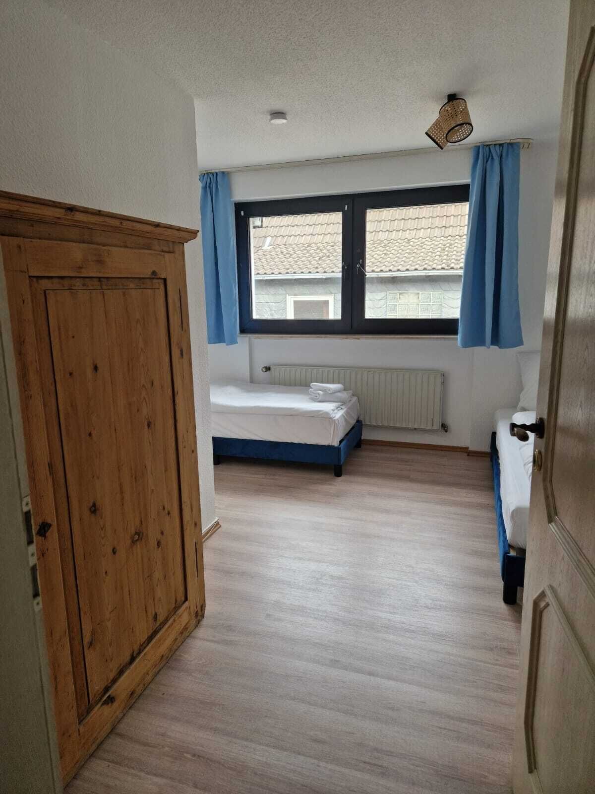 Une chambre avec une armoire en bois, un lit avec une literie blanche, une fenêtre avec des rideaux bleus et un radiateur placé sous la fenêtre.