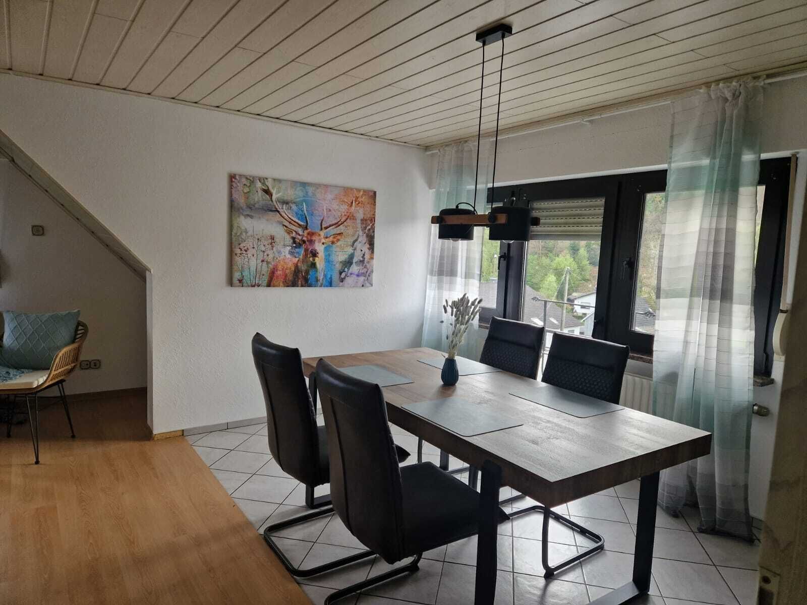 Un coin repas avec une table en bois, des chaises noires, un tableau au mur et une fenêtre avec des rideaux.
