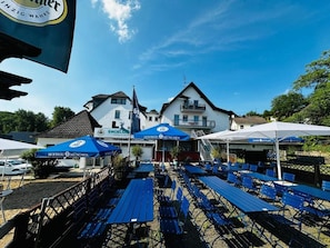 An outdoor seating area with tables and umbrellas, a building with a sign, and a clear sky. - Hotel Engelskirchen (Engelskirchen)