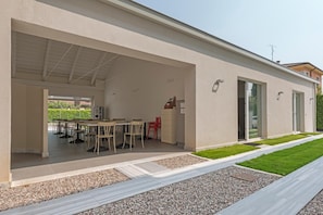 A modern building with a covered outdoor area featuring a dining table and chairs, a kitchen area, and a well-maintained lawn. - Garda Guest House (Lazise)