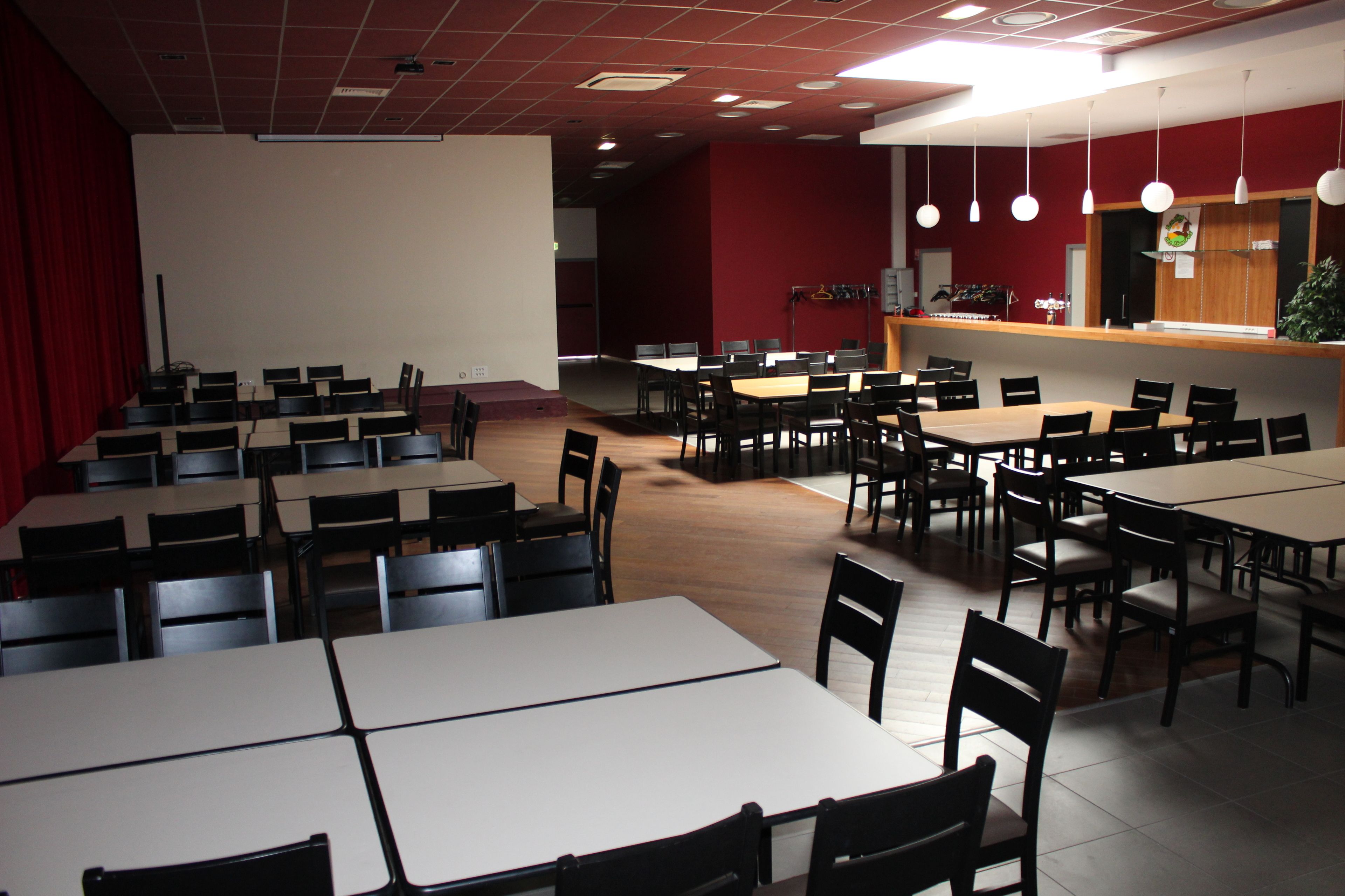 Un amplio comedor interior con varias mesas y sillas, un escenario con cortinas y una barra con una planta.