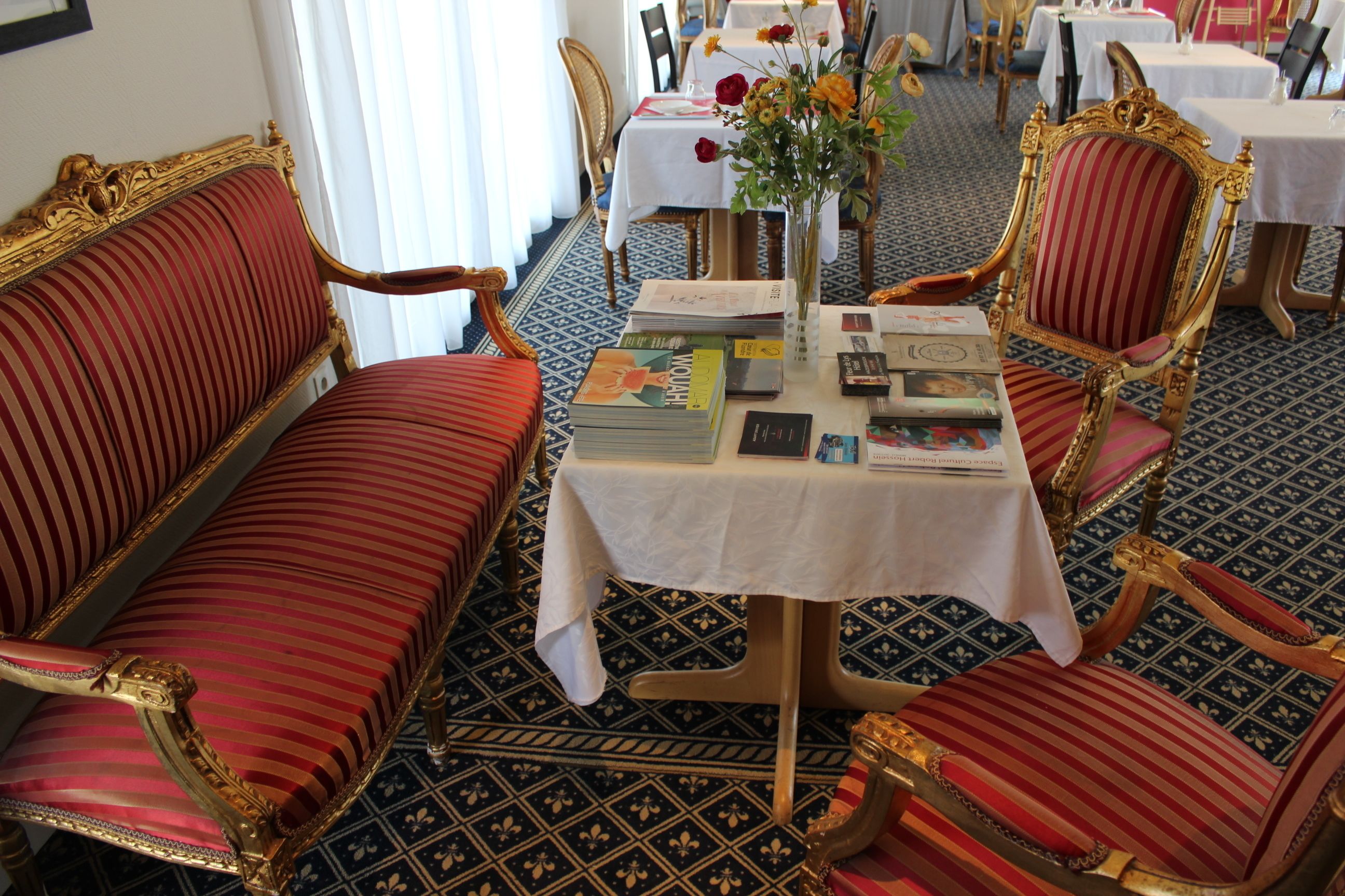 A room with ornate red upholstered chairs, a table with books and a vase of flowers, and patterned floor tiles.