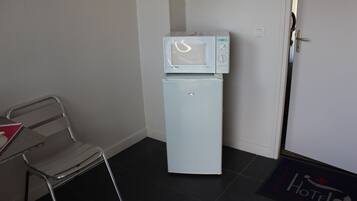 Una kitchenette compacta con microondas y refrigerador, una silla metálica y un felpudo de hotel.