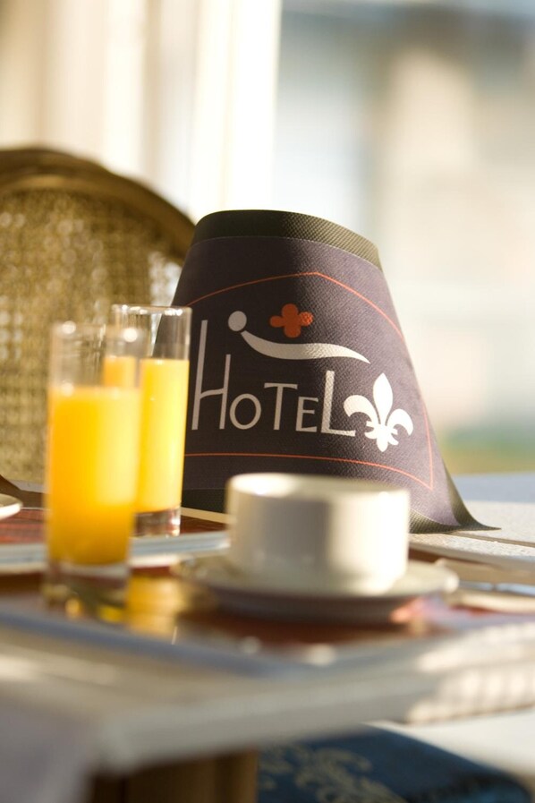 A hotel hat with the word "HOTEL" and a fleur-de-lis symbol, a table with a glass of orange juice, and a white cup. - Hôtel Fleur De Lys (Hazebrouck)