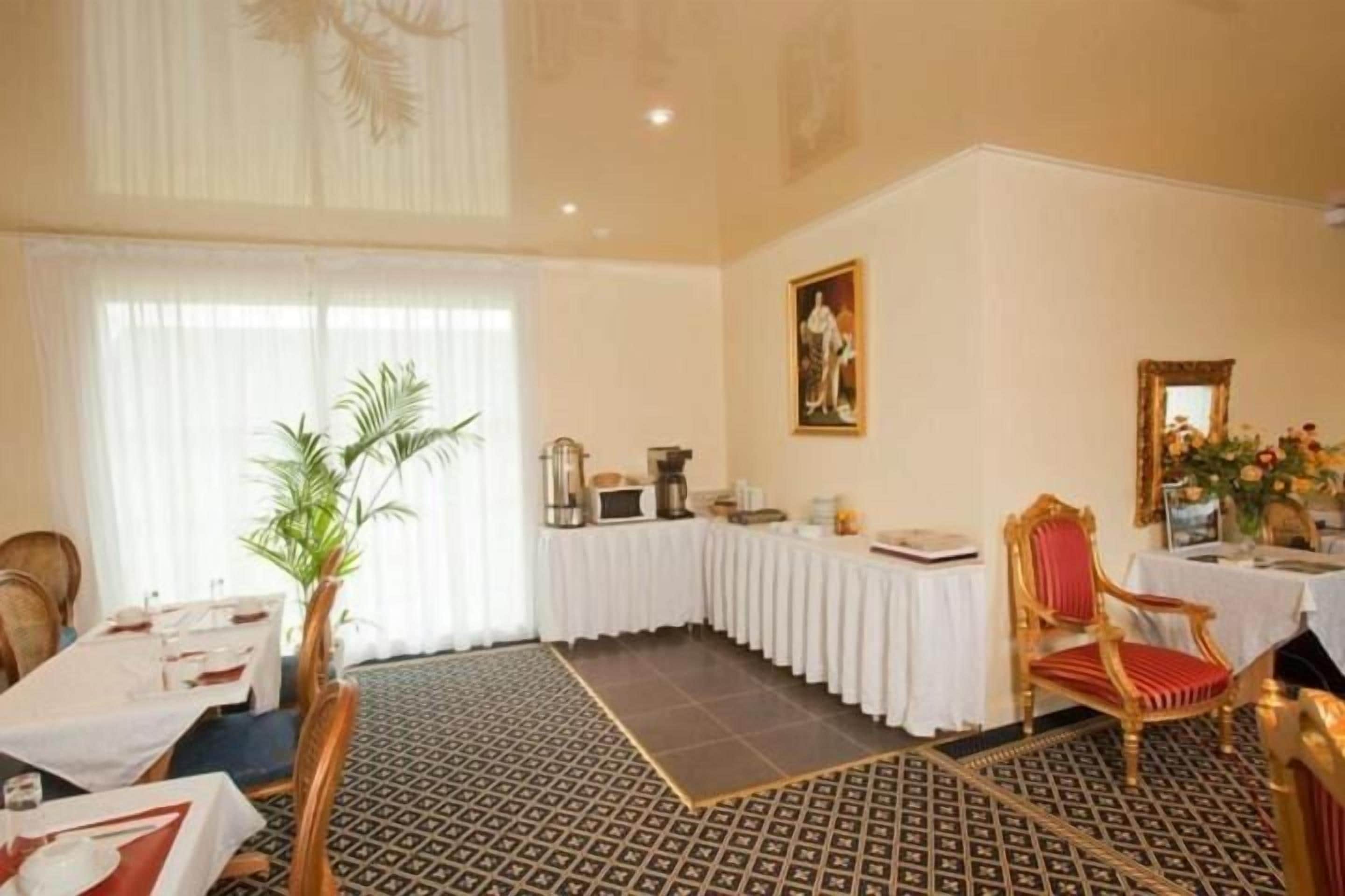 A hotel room with a dining area, a large window with sheer curtains, a painting on the wall, and a patterned floor.