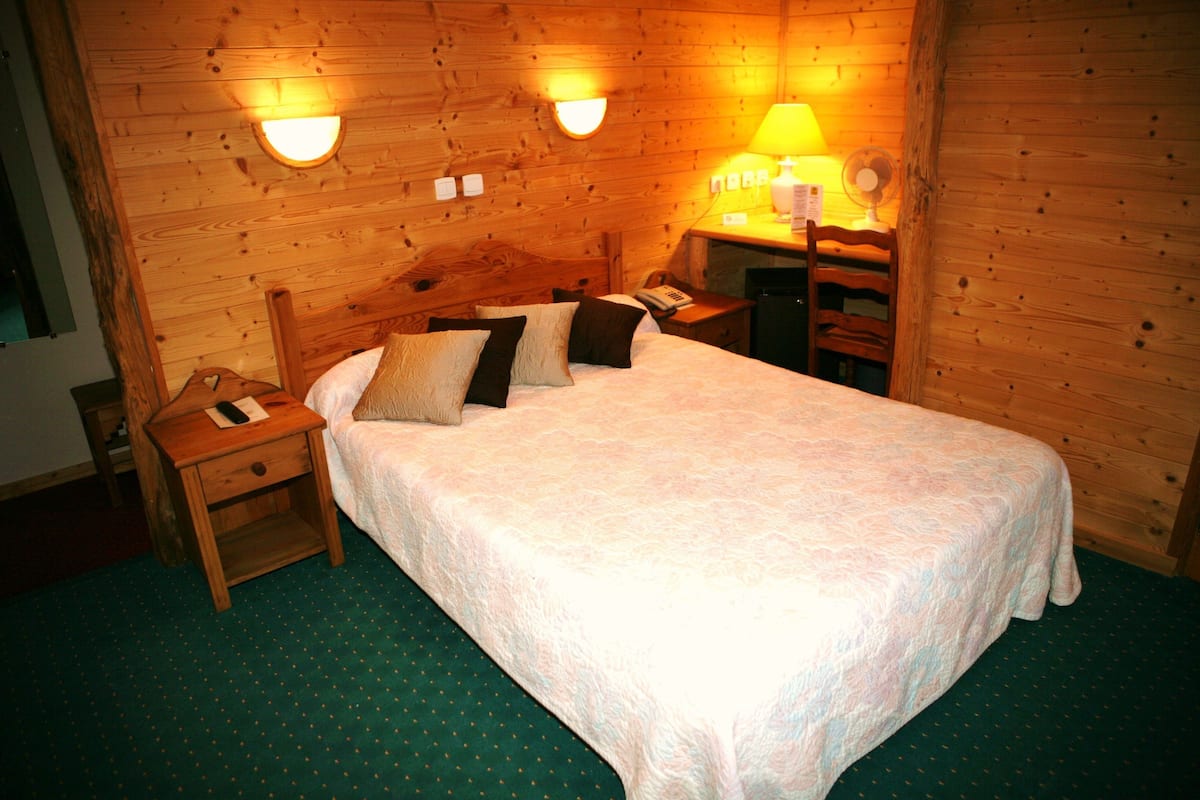 Een houten kamer met een bed, nachtkastje en een bureau met lamp en ventilator.