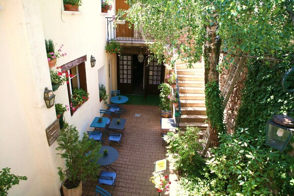 Un patio exterior con mesas y sillas, rodeado de edificios con plantas en macetas y una escalera de madera.