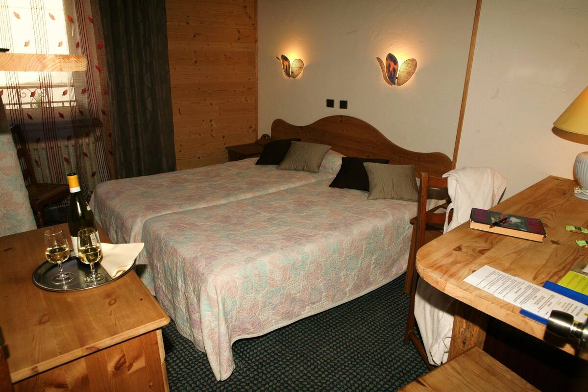 Een tweepersoonsbed met een geborduurd sprei, twee nachtkastjes, een houten bureau met een lamp en een fles wijn aan de linkerkant.