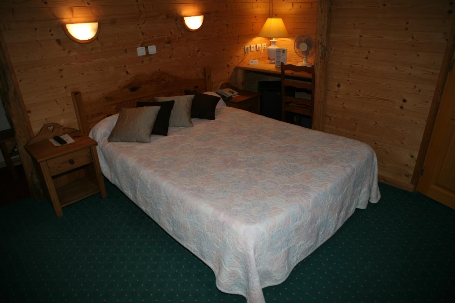 Een houten kamer met een bed, nachtkastje, lamp en wandlampen.