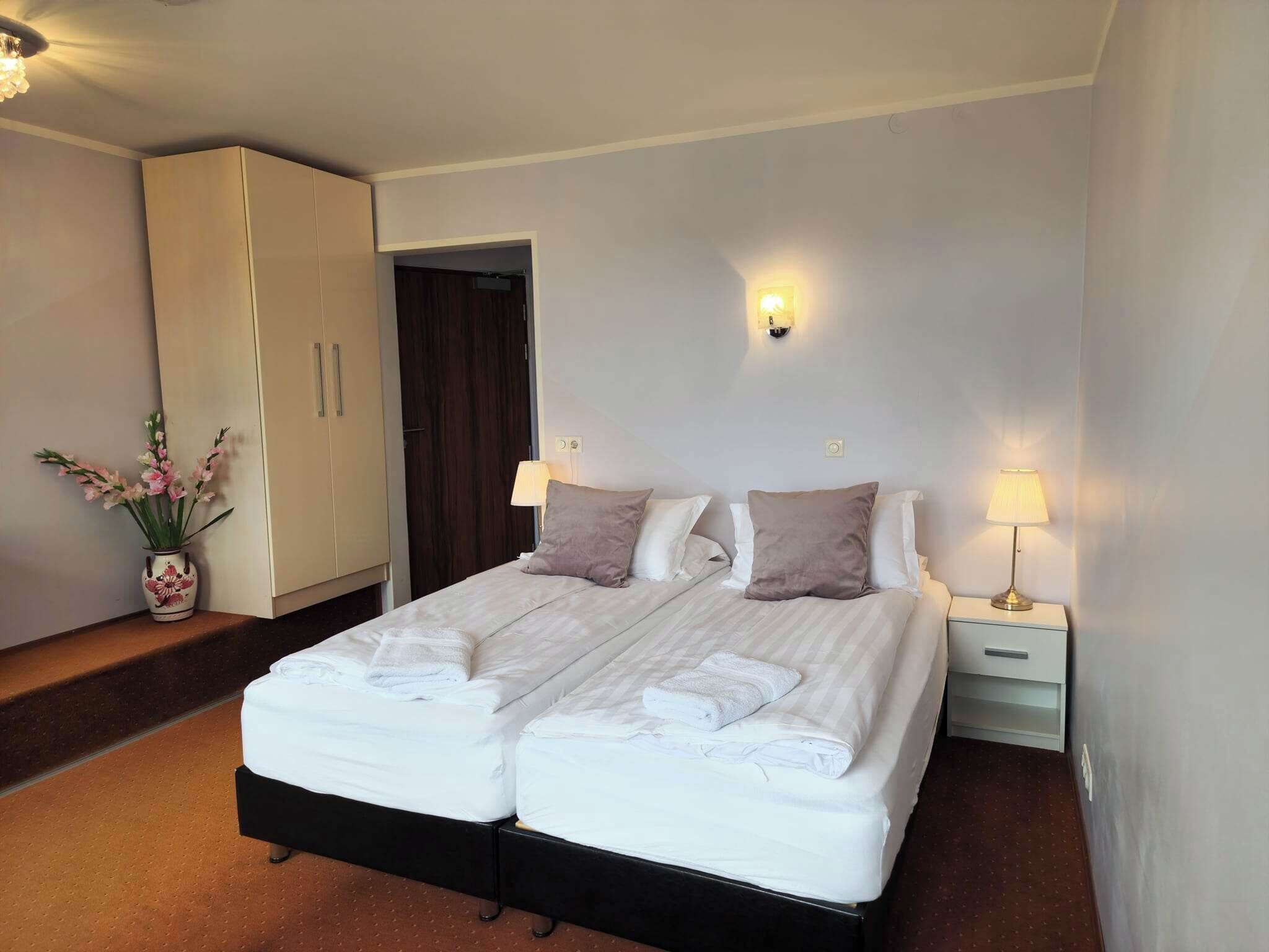 A double bed with white linens, two bedside lamps, a nightstand, a vase with flowers, and a wardrobe.
