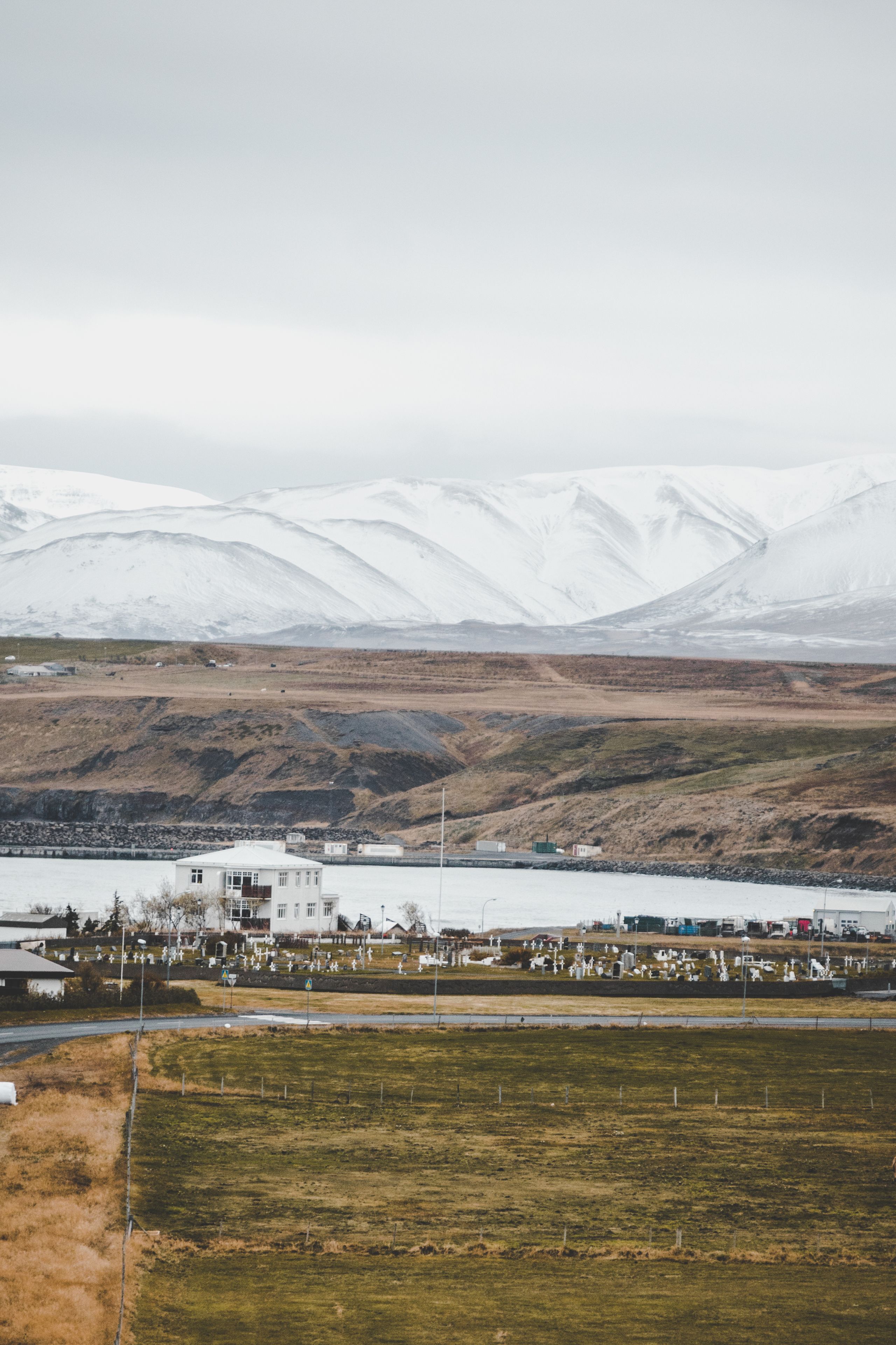 A rural landscape with a small settlement, a body of water, and snow-capped mountains in the background.
