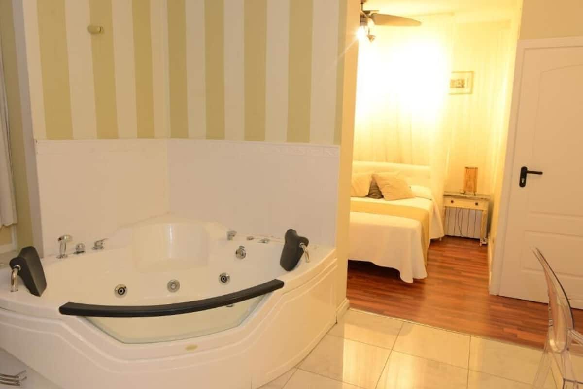 Une salle de bain avec une grande baignoire, un espace douche et une chambre séparée comprenant un lit et une table de chevet.