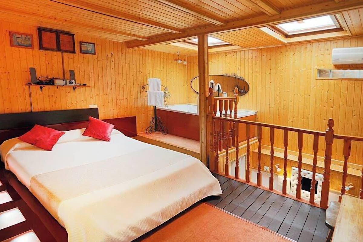 Une chambre située dans un chalet en bois, comprenant un lit, une petite table, une table de chevet et une salle de bain avec un lavabo et un miroir, ainsi qu’un balcon doté d’une balustrade en bois.