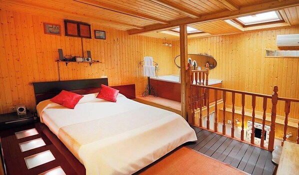 A wooden cabin bedroom with a bed, a small table, a nightstand, a bathroom with a sink and mirror, and a balcony with a wooden railing. - La Mansión de Navalcarnero (Navalcarnero)