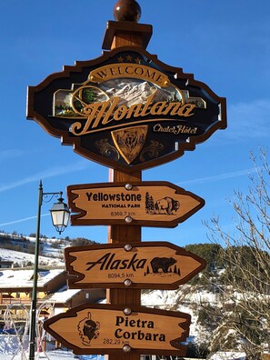 Un cartel de madera con la inscripción 'Welcome to Montana' en la parte superior, seguido de indicaciones hacia el Parque Nacional Yellowstone, Alaska y Pietra Corbora, con sus respectivas distancias.