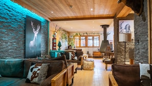A cozy living room with a wooden ceiling, a large deer antler decoration, a fireplace, and a stone wall. - Montana Chalet Hôtel Spa (Enchastrayes)