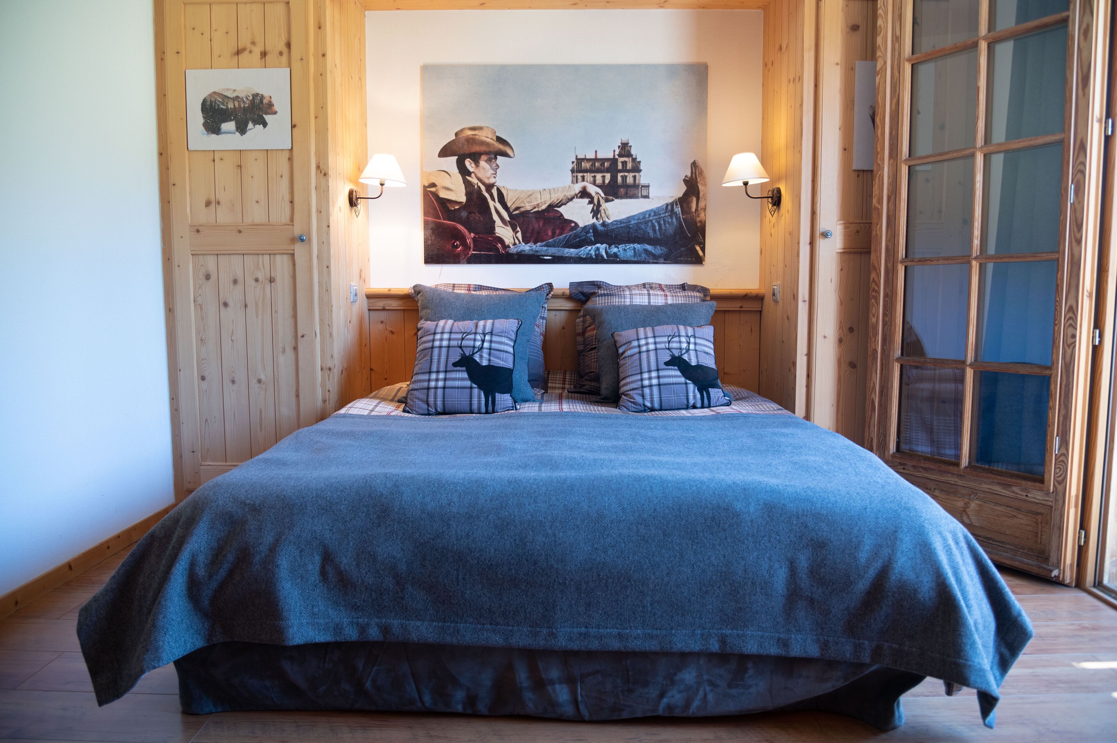 A bed with a blue blanket and plaid pillows, a wooden door, and a painting of a cowboy on the wall.