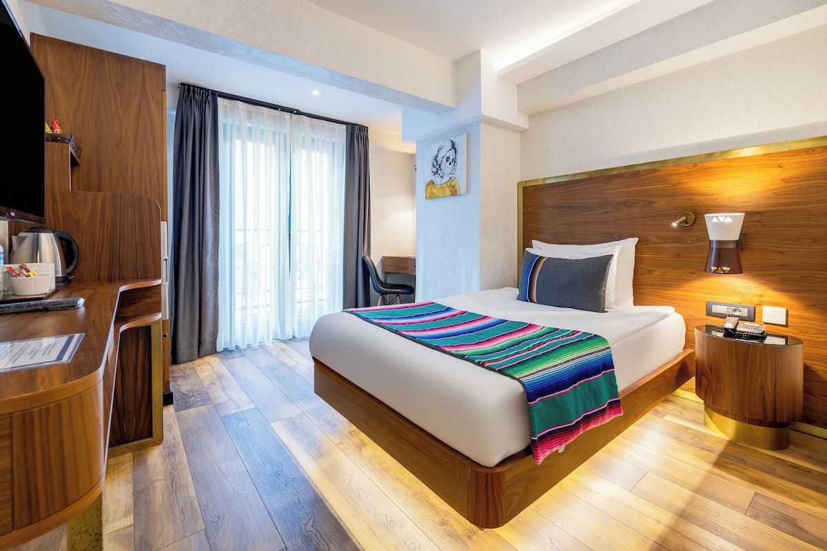 A modern hotel room with a wooden bed, a desk with a kettle, a chair, and a large window with curtains.