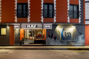 Fachada de hotel con un llamativo letrero 'HAY HOTEL', una entrada moderna con escalones y un mural decorativo con motivos de hojas a la derecha.