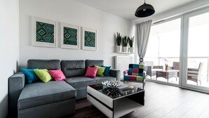 Una sala de estar moderna con un sofá gris, cojines coloridos, una mesa de centro de vidrio y grandes ventanales con vistas al exterior.