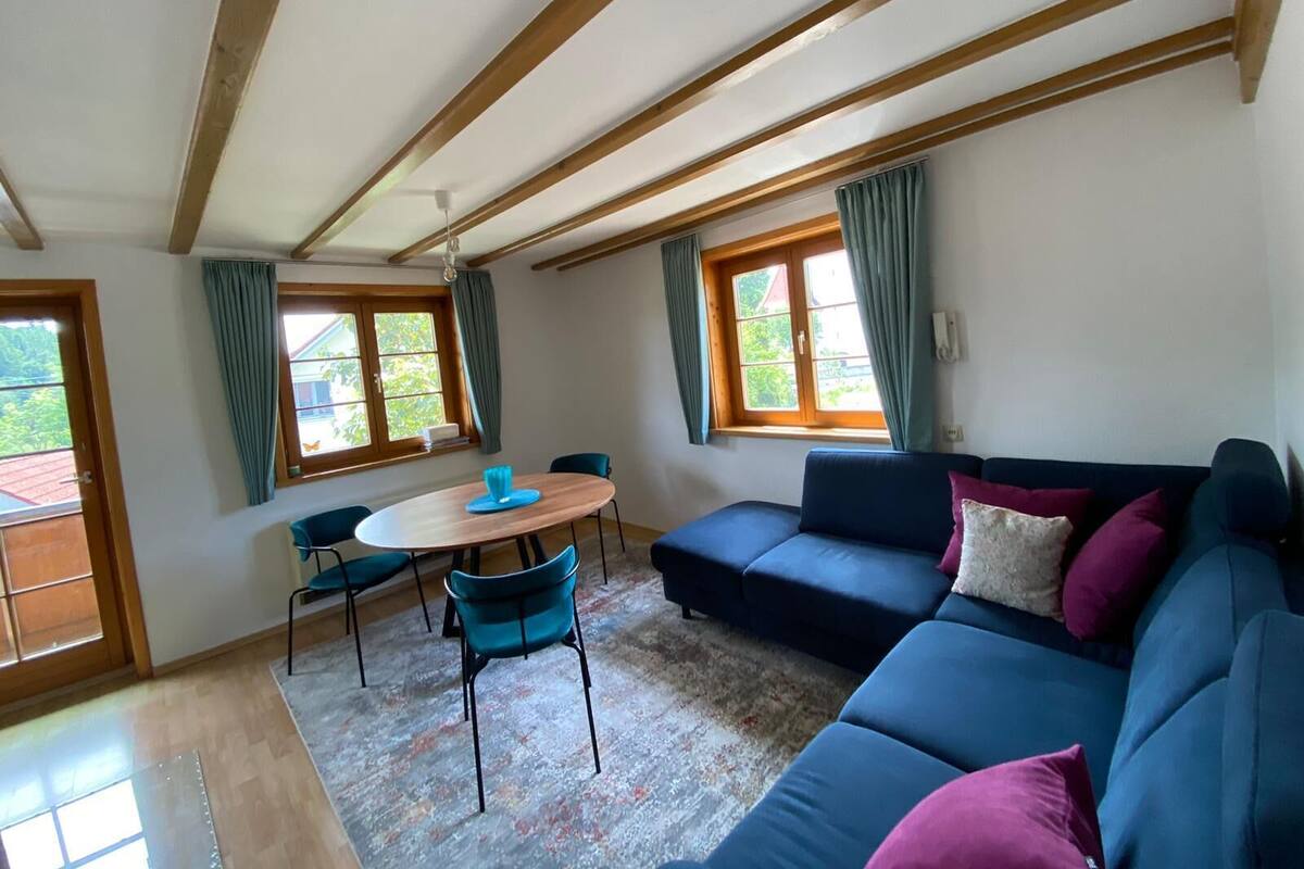 Une pièce avec un plafond en bois, un canapé bleu, une table ronde avec des chaises et de grandes fenêtres avec des rideaux.