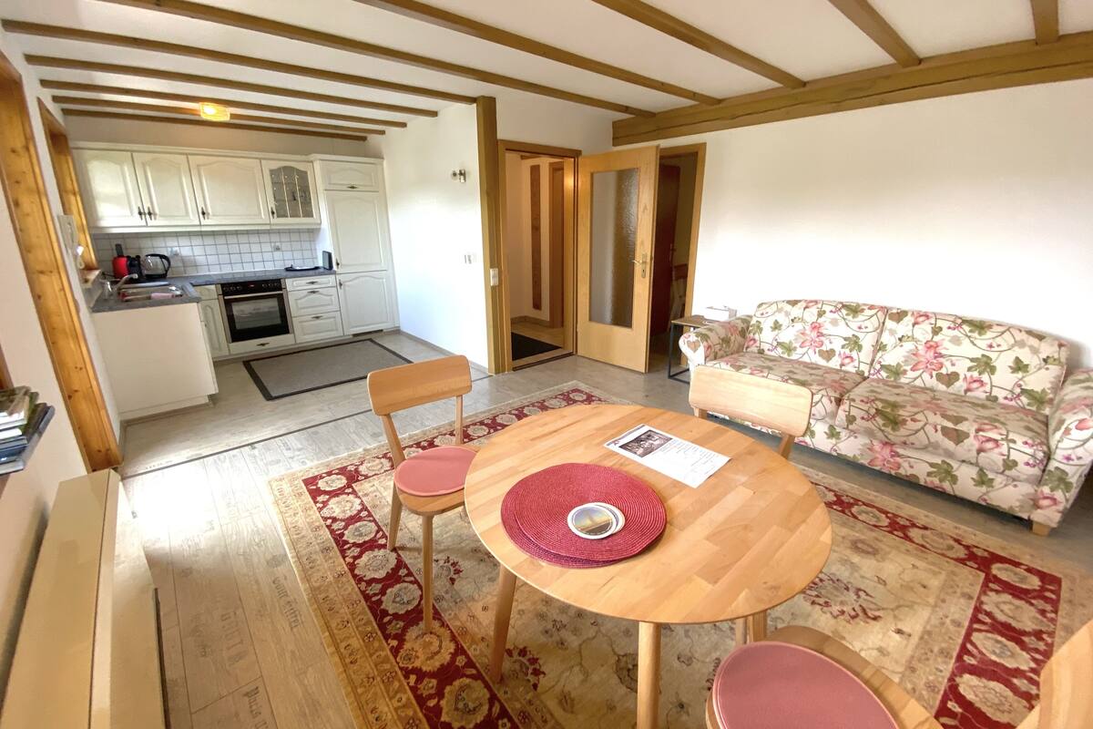Un salon chaleureux avec une table en bois, deux chaises, un canapé à motifs floraux et une cuisine en arrière-plan.