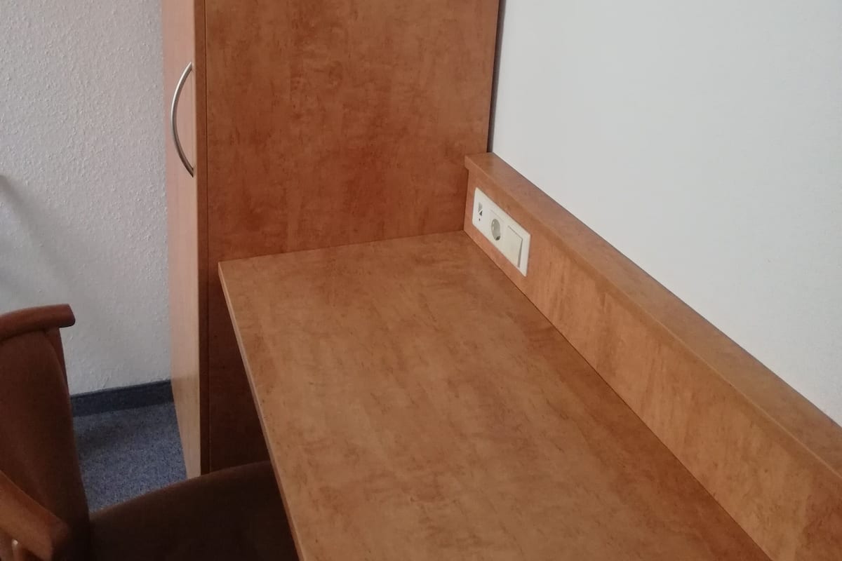 A small, wooden desk with a chair, a wall socket, and a door.