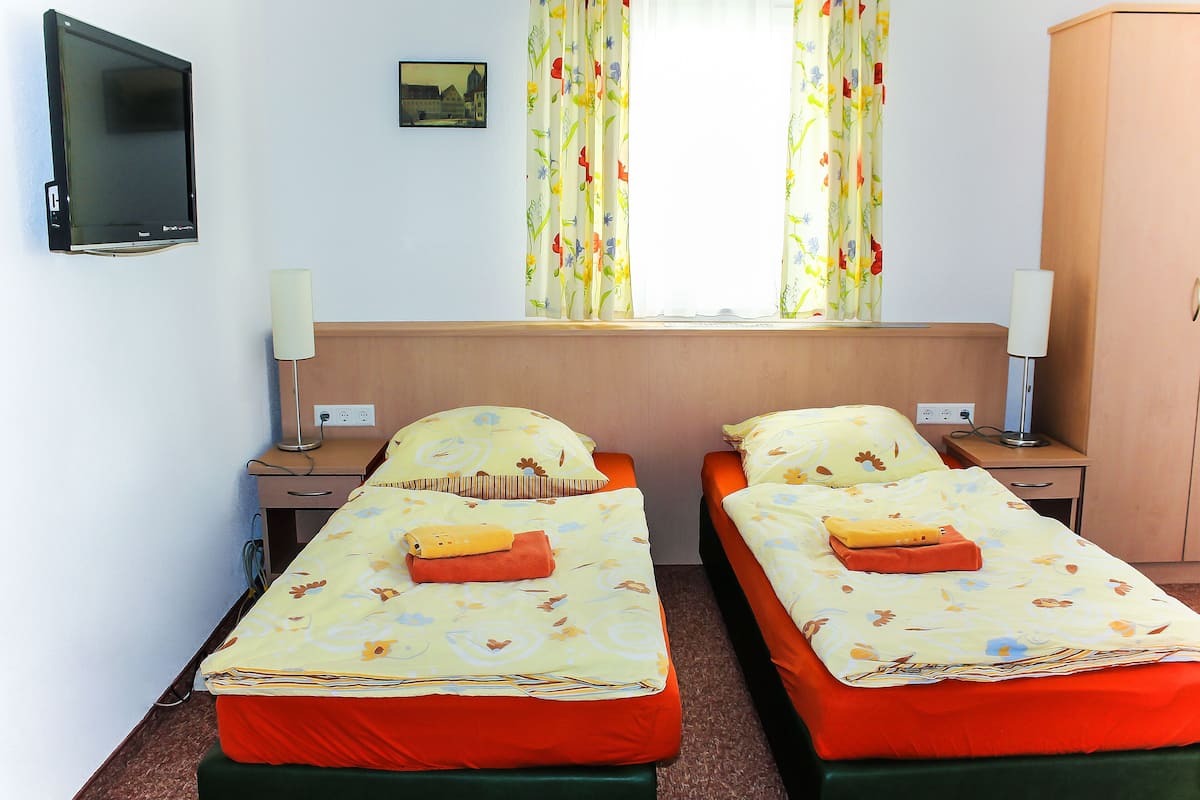 A hotel room with two single beds, a TV mounted on the wall, a window with floral curtains, bedside lamps, and a wooden wardrobe.