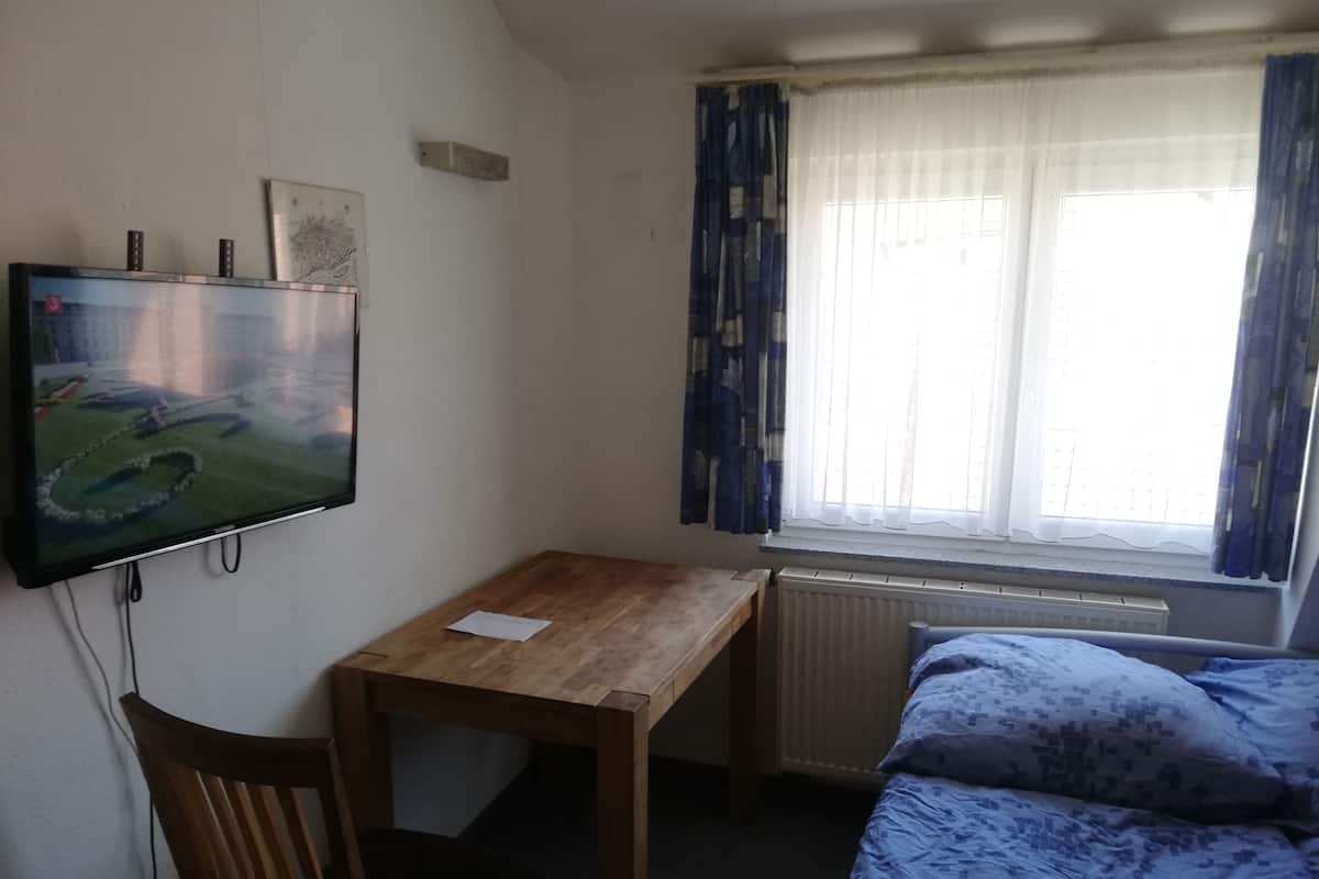 A room with a wooden desk, a television mounted on the wall, a window with curtains, and a bed with patterned bedding.