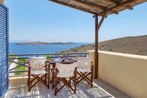 A balcony with a wooden table and two chairs, overlooking a coastal landscape with a lighthouse in the distance. - Sunrise Beach Suites (Syros)