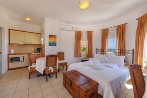 A hotel room with a bed, a wooden chest, a dining table with chairs, a kitchen area, and a view of the sea through the window. - Sunrise Beach Suites (Syros)