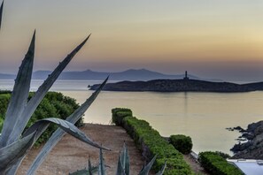 A coastal landscape with a clear view of the sea, a lighthouse, and a pathway leading to the water. - Sunrise Beach Suites (Syros)