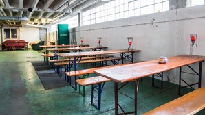 A spacious indoor area with multiple wooden tables and benches, a green floor, and a large window providing natural light. - Boot Camp Central - Hostel (Friedrichshafen)