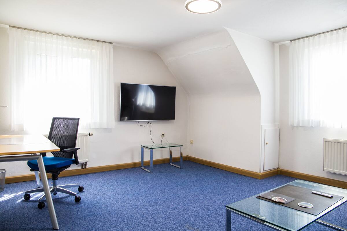 Una sala de conferencias con una gran pantalla plana, una silla de oficina azul, una mesa de vidrio y piso cubierto de alfombra azul.