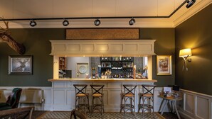 Un espace bar avec un comptoir blanc, trois tabourets de bar, une étagère remplie de bouteilles et un luminaire fixé au mur.