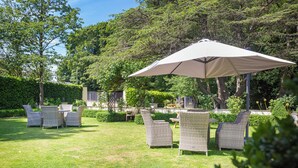 Un espace extérieur aménagé avec des chaises en osier et un grand parasol, entouré d’une végétation luxuriante et d’arbres.