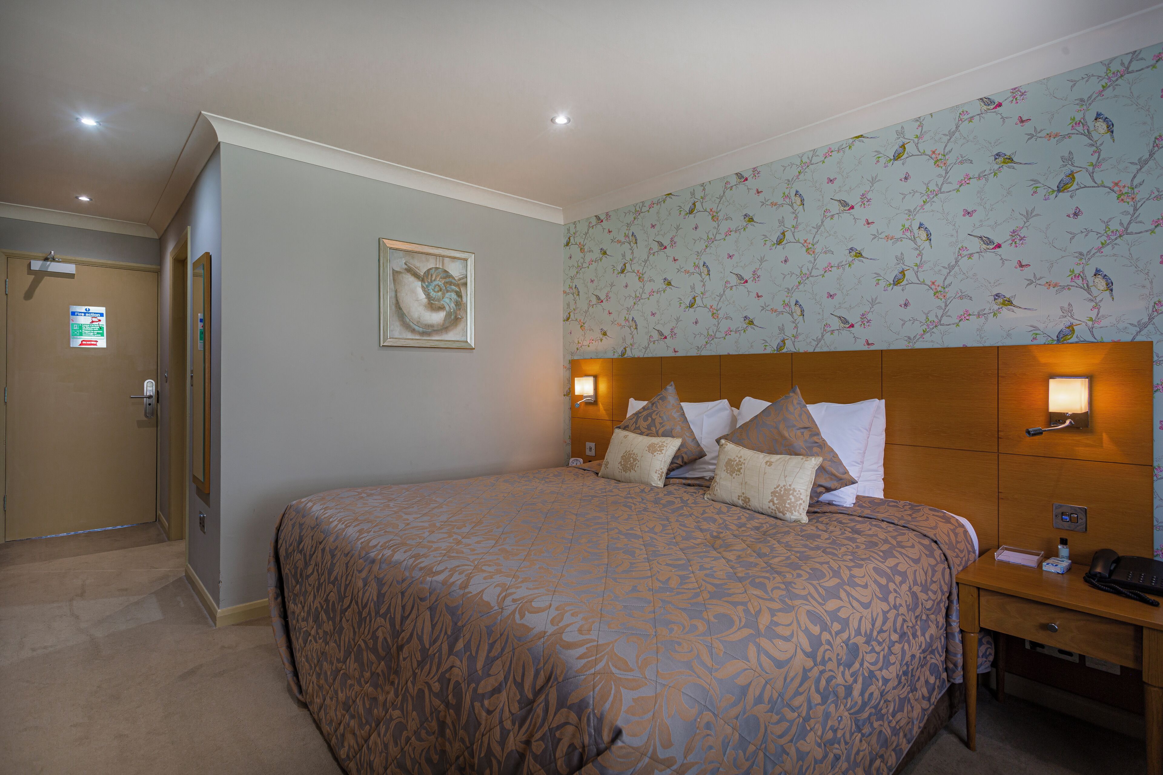 A hotel room with a large bed, a wooden headboard, a nightstand, and a wall-mounted lamp.
