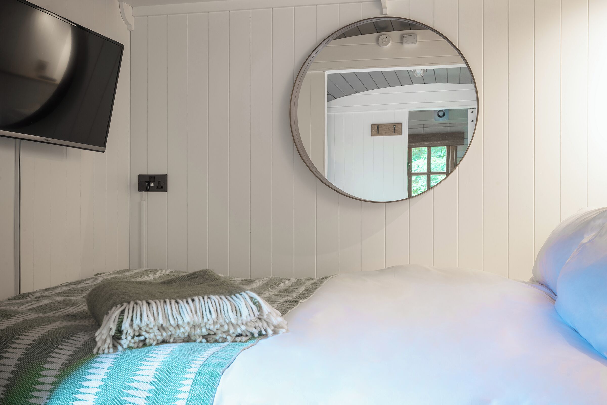 A bedroom with a bed, a round mirror, and a television on the wall.