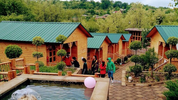 A wooden lodge with a pond, surrounded by greenery and a hilly landscape.