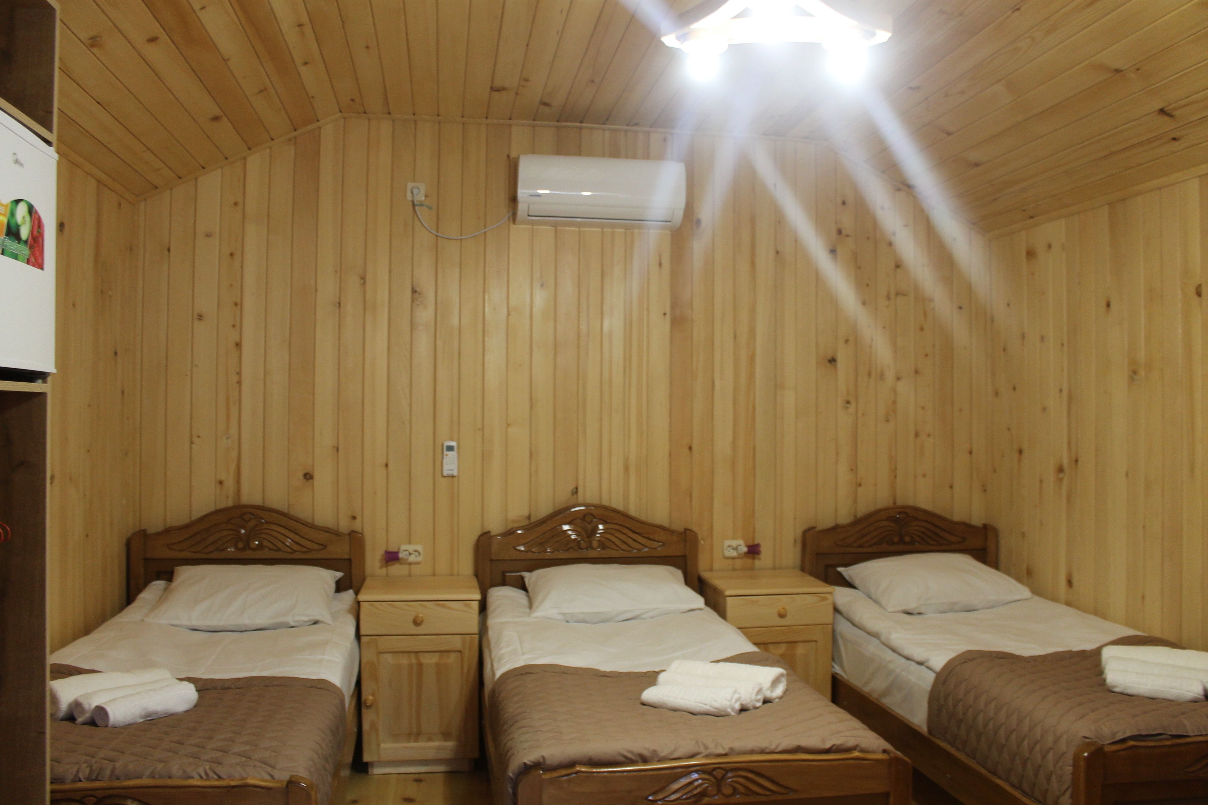 Quarto com painéis de madeira e três camas individuais, cada uma com cabeceira de madeira, mesa de cabeceira e criadeira. Um aparelho de ar condicionado está fixado na parede acima das camas.