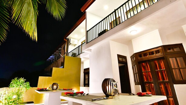 A well-lit outdoor dining area with a table set for two, surrounded by palm trees and a modern building with a balcony.
