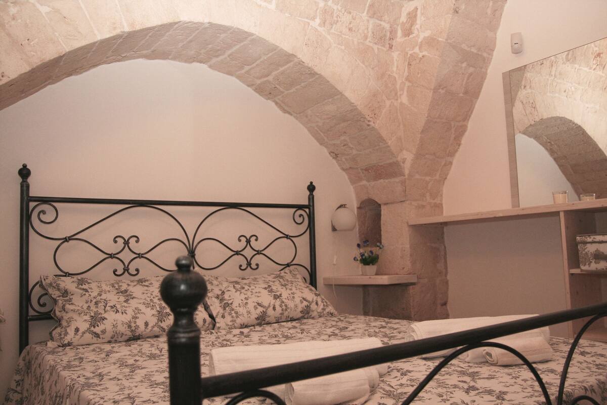 A bedroom with a bed, a nightstand, and a mirror. The room has stone arches on the ceiling and a brick wall.
