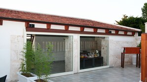 Un patio con suelo de baldosas, pared blanca y techo de tejas rojas. Se observan dos plantas en macetas y una silla de madera.