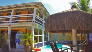 Un espace piscine doté d’une structure à toit de chaume, de chaises longues et d’un bâtiment avec un balcon.