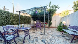 Eine Terrasse im Freien mit Steinboden, lilafarbener Metallmöbel, einem überdachten Sitzbereich und einer grünen Pergola.