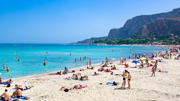 Ein belebter Strand mit klarem, blauem Wasser, Menschen, die sich sonnen, und Booten.