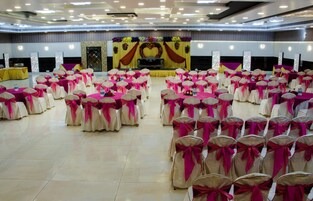Un salón de banquetes con filas de mesas y sillas, un escenario con decoraciones florales y un suelo de baldosas.