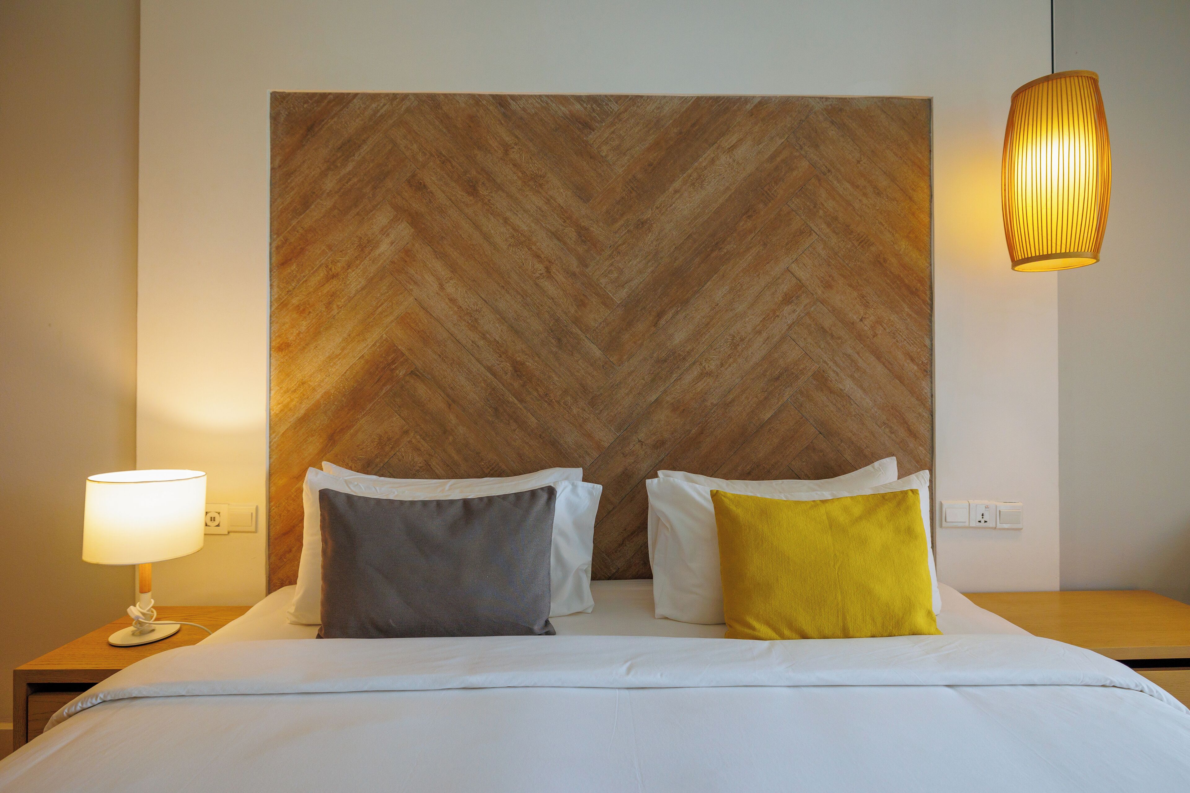 Ein Hotelzimmer mit Holzboden-Paneelen am Kopfteil des Bettes, weißer Bettwäsche, zwei Kissen (ein graues, ein gelbes), einer Nachttischlampe und einer Pendelleuchte.