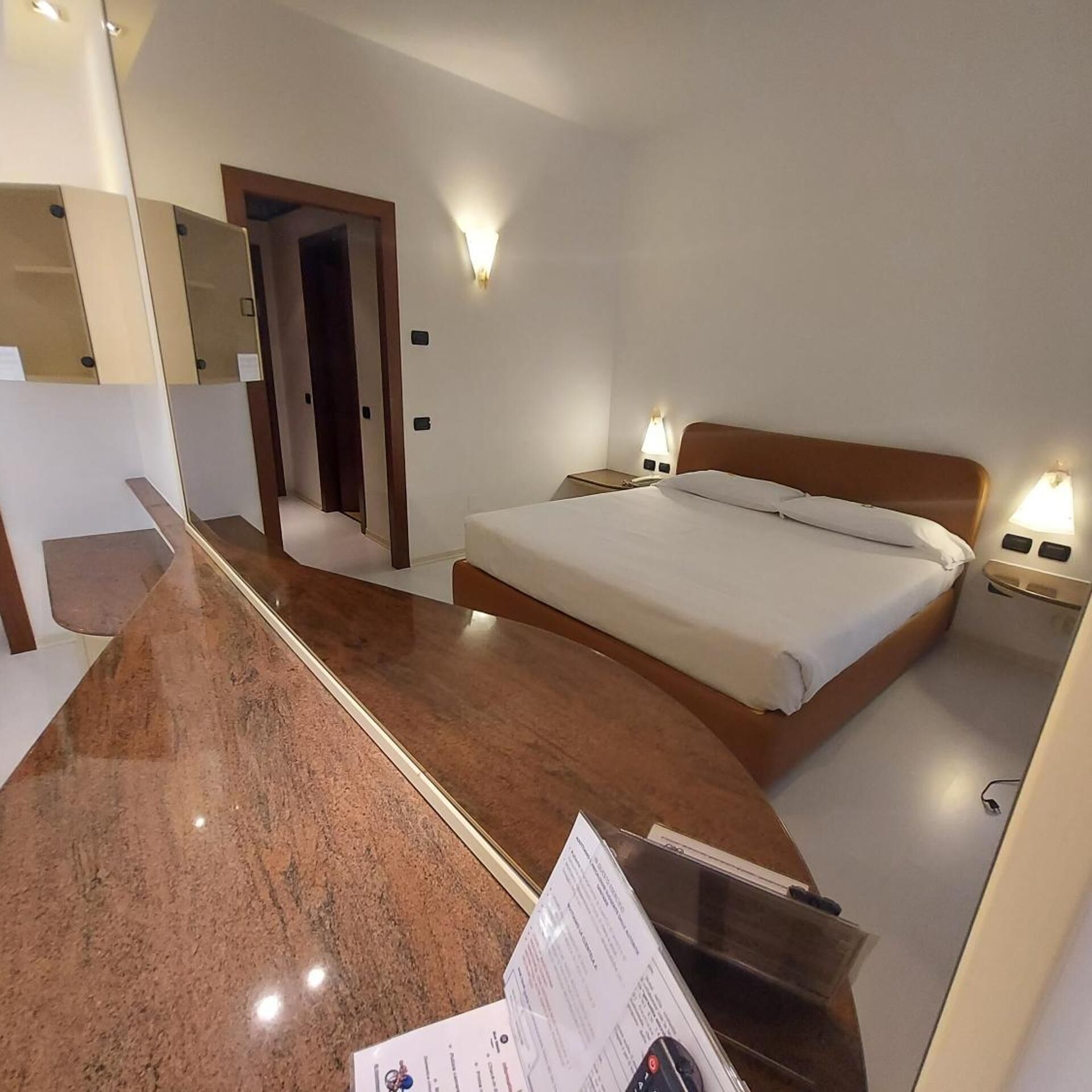 A hotel room with a bed, bedside tables, a mirror, and a reception desk.
