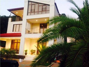 A two-story building with balconies, a covered entrance, and a landscaped garden with palm trees. - Odissea's (Batumi)